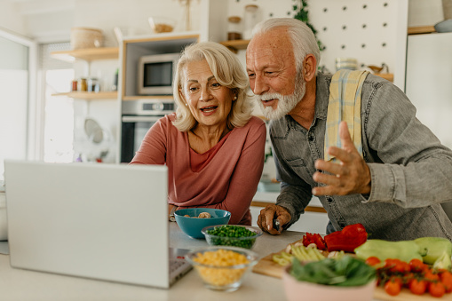 Cheerful senior couple preparing a healthy meal using an online recipe. Senior man cutting vegetables while his wife is checking a recipe online on the laptop.