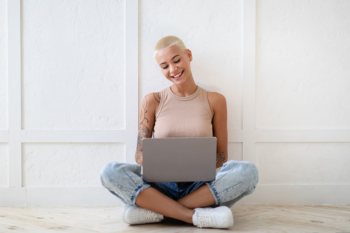 Happy young woman sitting on floor with modern laptop on white wall background, working online or surfing on Internet, free space