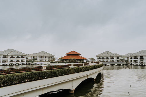 A wide shot of the cityscape view of Hanoi in Vietnam, depicting multiple white minimalist modern western style houses and a traditional vietnamese residential building or ancient architecture with a red top in the middle, in front of a bridge on the water of the river or sea, in a overcast weather or gloomy day.