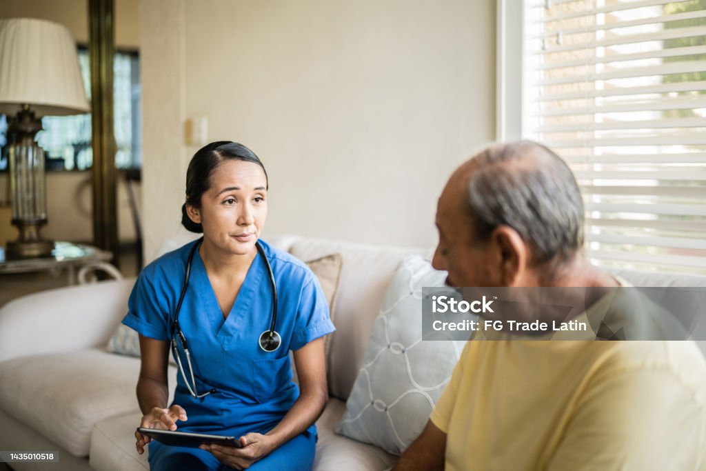 Mid adult nurse using digital tablet while talking to a senior man in a consultation in the living room of a nursing home Home Caregiver Stock Photo