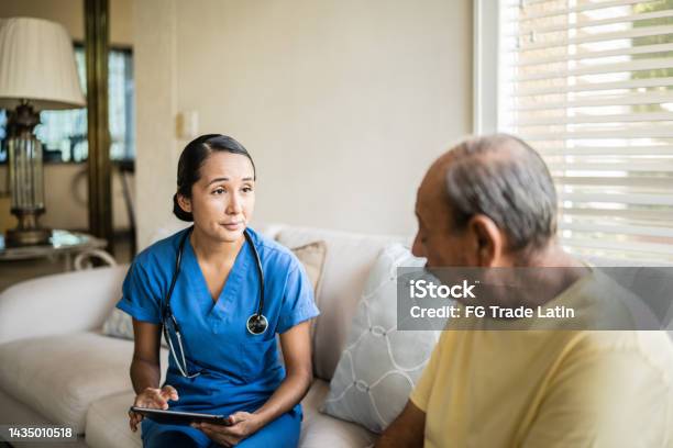 Mid Adult Nurse Using Digital Tablet While Talking To A Senior Man In A Consultation In The Living Room Of A Nursing Home Stock Photo - Download Image Now