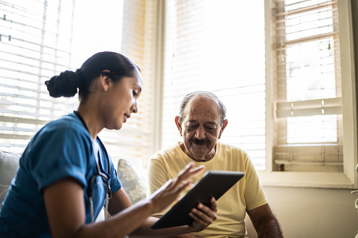 Mid adult nurse using digital tablet while talking to a senior man in a consultation in the living room of a nursing home