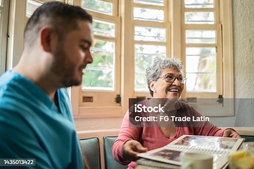 istock Senior woman and mid adult nurse looking at photo album in the dining room in a nursing home 1435013422