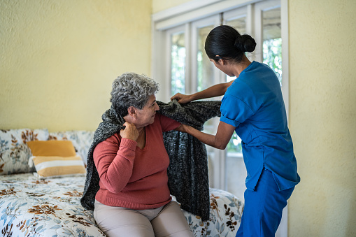Mid adult nurse helping senior woman dress shirt in bedroom at nursing home