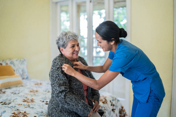 infermiera adulta di metà che aiuta la camicia della donna anziana in camera da letto alla casa di cura - assisted living foto e immagini stock