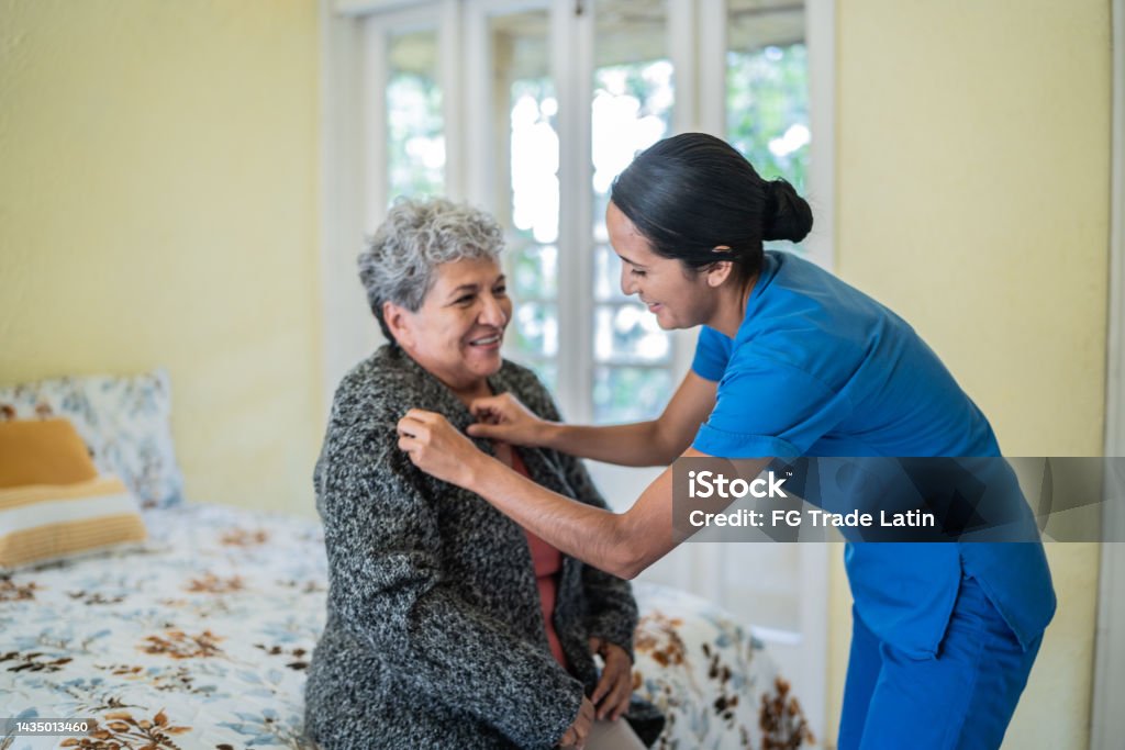 Mid adult nurse helping senior woman dress shirt in bedroom at nursing home Assisted Living Stock Photo