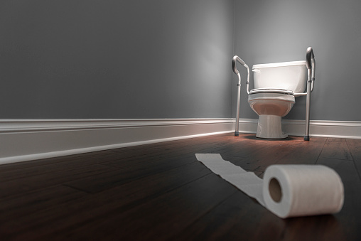 An elongated white toilet with retrofit safety grab bars with runaway roll of toilet paper in the foreground. The toilet is highlighted with a spot light coming from above. This sparse room has gray walls and a dark hardwood floor.