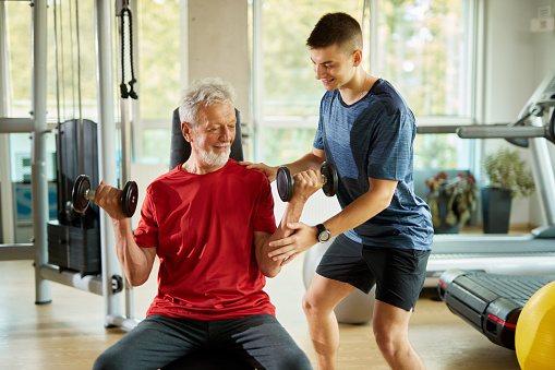 Senior man exercising with personal trainer using dumbbells at the gym