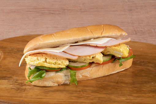 fresh submarine sandwiches with ham, cheese, bacon, tomatoes, lettuce, cucumbers and onions on dark wooden background
