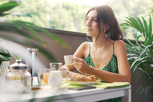 Young happy woman sitting in the balcony and having breakfast, she is drinking coffee