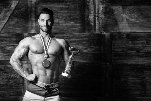 Bodybuilder Competitor Showing His Winning Medal Against a Wooden Wall and Flexing Muscles While Wearing Sports Shorts - Male Fitness Competitor Showing His Winning Medal a Place for Your Text