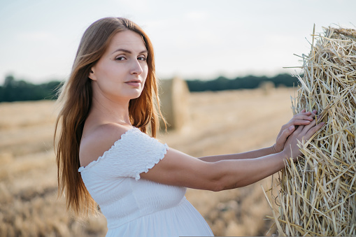 beautiful pregnant brunette in a white dress near the haystacks in the field at sunset. High quality photo
