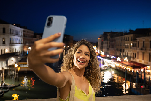 Happy female tourist taking a selfie at night in Venice using her cell phone - traveling concepts