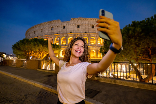 Happy travel influencer vlogging from the Coliseum in Rome at nighttime using her cell phone - travel concepts