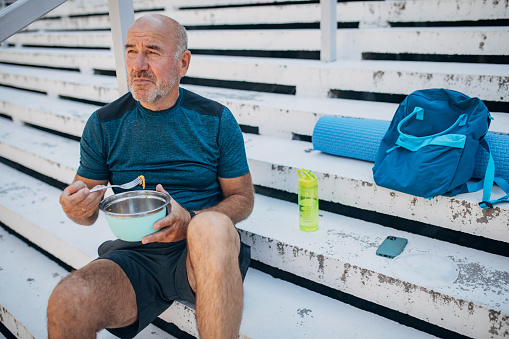One woman, mature male taking a break sitting on steps outdoors after exercising. He is eating a salad.