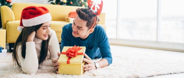 młoda szczęśliwa azjatka w kapeluszu świętego mikołaja ze swoim chłopakiem z prezentem świątecznym, leżąc na dywanie i patrząc w kamerę z choinką w tle. - giving christmas lifestyles holiday zdjęcia i obrazy z banku zdjęć