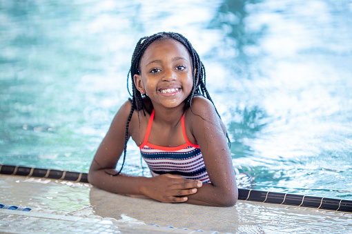 A sweet young girl of African decent poses at the side of a community pool for a portrait.  She is wearing a striped swim suit and is smiling with her wet hair slicked back.