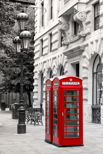 Bright red London phone boxes on a city street with lamp post, benches and trees in the background. Black and white with the telephone booths picked out in red.