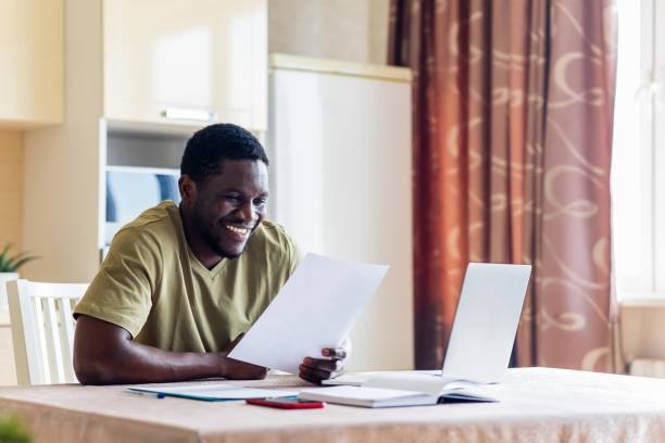 latynoski latynoski mężczyzna wyglądający na szczęśliwego, siedząc z laptopem i papierami w kuchni - african descent confidence african culture education zdjęcia i obrazy z banku zdjęć