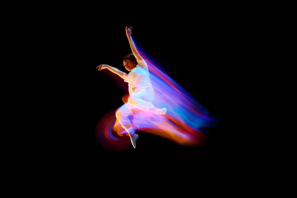 Dance in motion. Studio shot of flying, jumping dancer or gymnast performing tricks in the air over black background with mixed neon glowing rays Dance in motion. Studio shot of flying, jumping dancer or gymnast performing tricks in the air over black background with mixed neon glowing rays. Fantasy, cyberpunk, sport, fashion hyperdrive stock pictures, royalty-free photos & images
