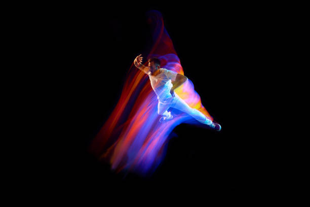 Dance in motion. Studio shot of flying, jumping dancer or gymnast performing tricks in the air over black background with mixed neon glowing rays Dance in motion. Studio shot of flying, jumping dancer or gymnast performing tricks in the air over black background with mixed neon glowing rays. Fantasy, cyberpunk, sport, fashion hyperdrive stock pictures, royalty-free photos & images