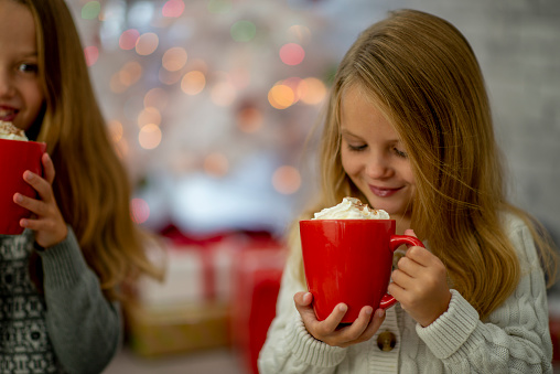 A sweet little blond haired girl holds up a cup of Hot Chocolate as she takes a sip on Christmas morning.  She is dressed casually and is smiling.