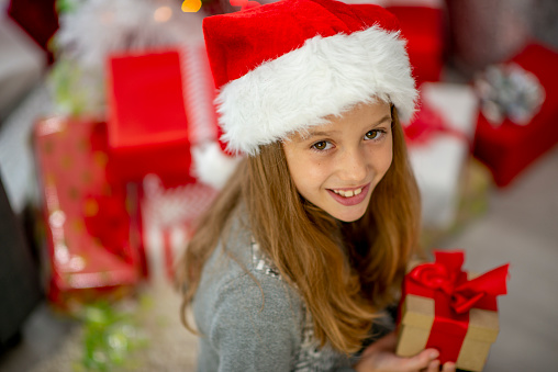 A sweet little blond haired girl with a Santa hat on, looks up to the camera as she holds a small gift box with a red bow.  She is dressed in a warm sweater and is seated in front of her Christmas tree.