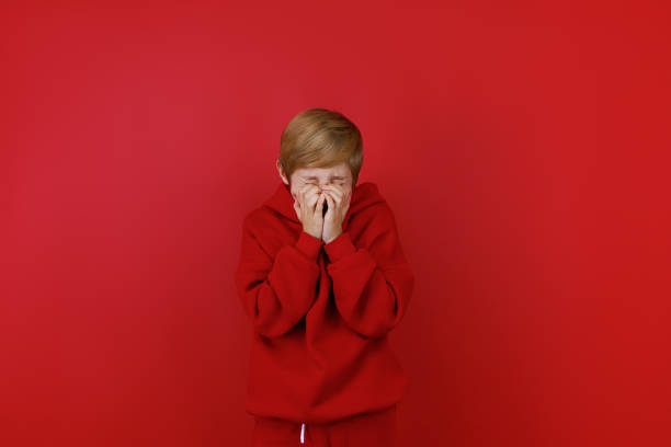 frightened girl in studio. she close her face. caucasian child stand afraid of something, looks at camera with one eye, isolated on red background