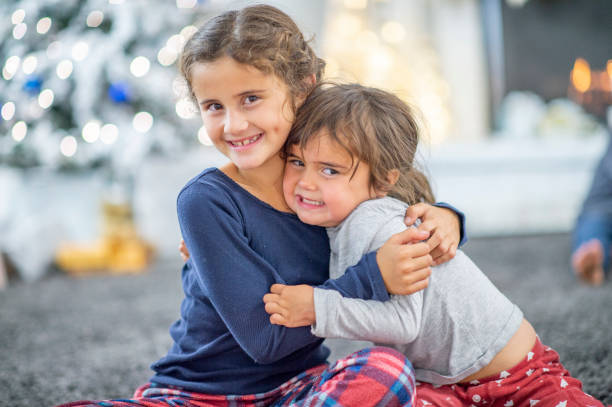 Two sweet sisters hug each other on their living room floor on Christmas morning. They are both in pajamas and their Christmas tree can be seen in the background as they enjoy the warmth of the fireplace.