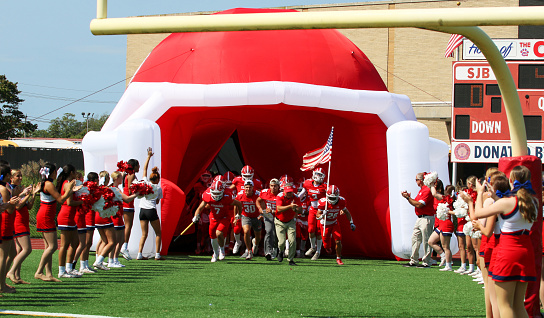 West Islip, New York, USA - 17 September 2022: High school football team entering trhe field through a blow up football helmet tunnel with cheerleaders in a line cheering.