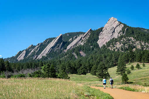 A hiking trail in Chautauqua Park below the Flatirons. The rock formations dominate the foothills of the Rocky Mountains above Boulder. Hikers are visible on the trail\nBoulder, Colorado, USA\n07/12/2022