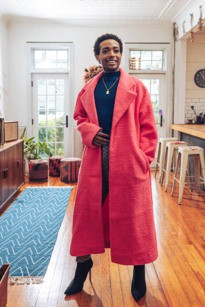 Home portrait of a young Non-Binary person. He/She is wearing a pink colored jacket and smiling to camera.