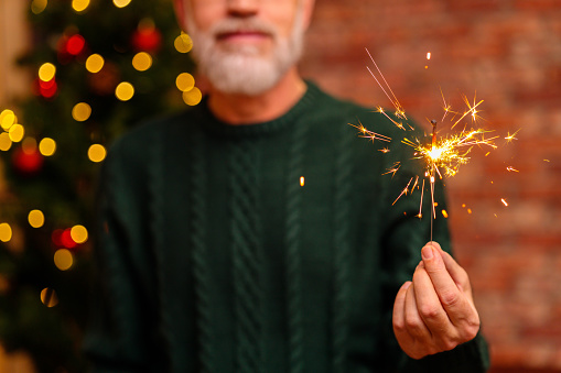 an elderly man in a green knitted with sparkler near the Christmas tree.