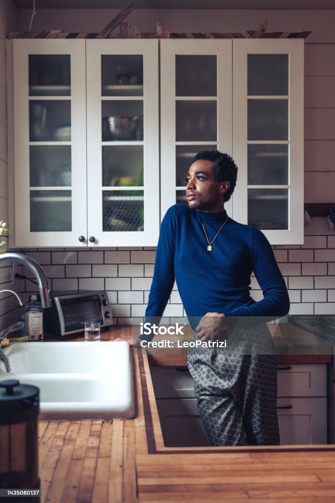 Non-binary person looking out of the window in the kitchen He/She staying in the kitchen and pensive looking out of the window. Bisexuality Stock Photo