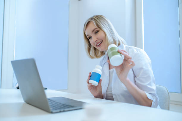 Smiling friendly general practitioner giving an online consultation Female physician holding plastic vitamin bottles in the hands and looking at the laptop screen Online Dermatology Courses stock pictures, royalty-free photos & images