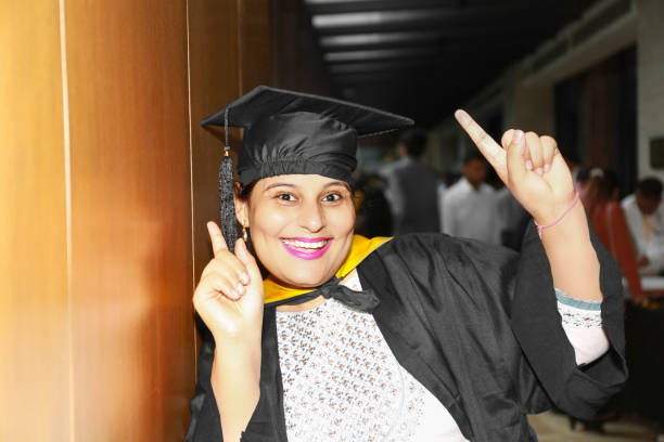 Young female of Indian ethnicity with toothy smile wearing graduation cap and gown portrait during convocation day in university.