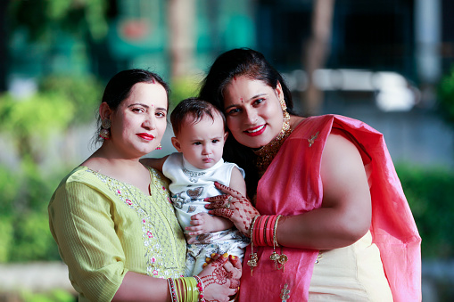 Two Cheerful women of Indian ethnicity standing outdoor in nature carrying baby child in hands.