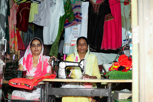 Two female tailor of Indian ethnicity stitching garment at home in workshop portrait together at home.