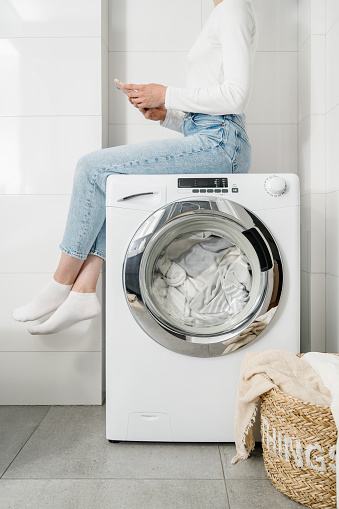 female housewife sitting with mobile phone on automatic washing machine with bed linen inside and closed door, wicker basket with clothes on floor in bathroom, cropped shot