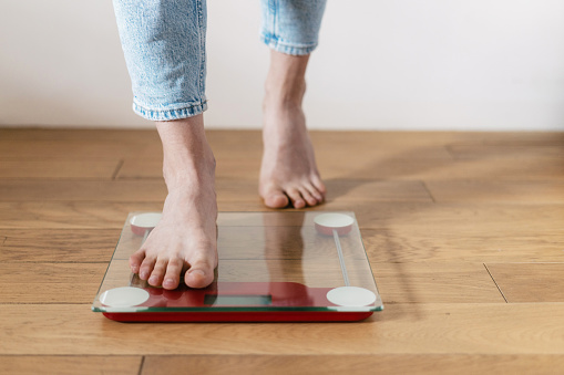 Cropped shot of young barefoot woman in casual blue jeans step on the floor scales to check her weight at home. Concept of bmi control and keeping the body in shape