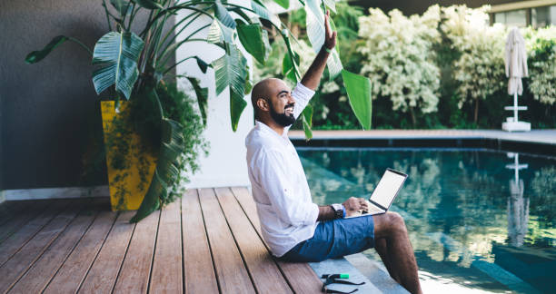 istock Cheerful Turkish digital nomad with mockup laptop computer waving while resting at pool terrace in own villa courtyard, successful male freelancer with blank netbook enjoying touristic job lifestyle 1435089816