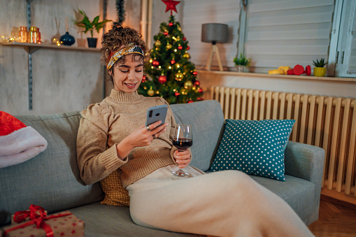 Young cheerful woman enjoying at home in a cozy Christmas atmosphere while using mobile phone