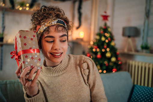 Happy young woman holding a Christmas gift near her ear, wondering what's inside