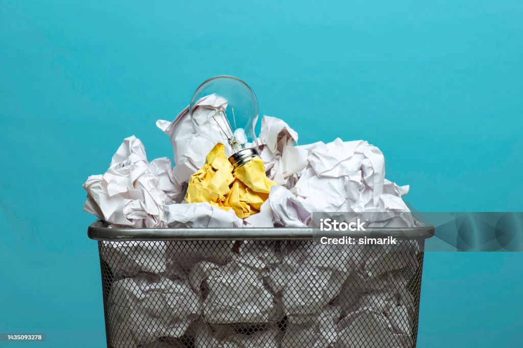 Solution Light bulb on a yellow crumpled paper ball among many white crumpled paper balls in a wastepaper basket on blue colored background. Ideas Stock Photo