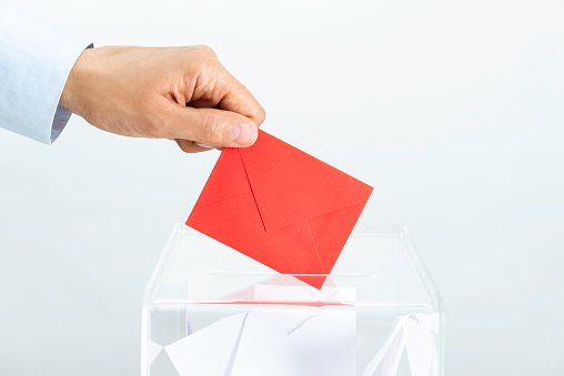 Human hand is inserting red envelope into ballot box in front of white background.