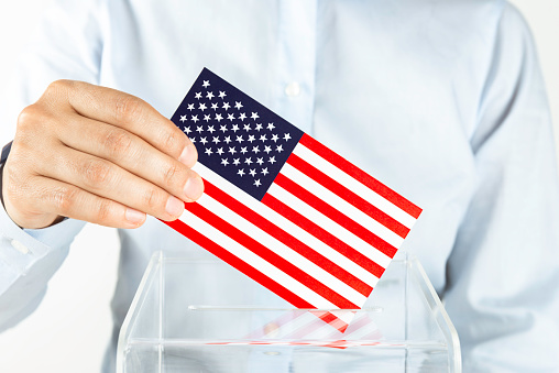 Human hand is inserting USA flag into ballot box. Representing voting in United States.