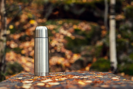 Steel thermos with delicious hot tea in the autumn forest on an old wooden table with fallen leaves in focus. The background is blurred.