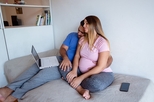 pregnant venezuelan caucasian heterosexual couple sitting on bed using laptop, surfing internet watching series online, enjoying day off at home