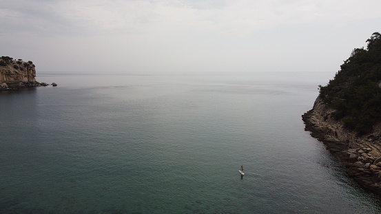 A person on a stand-up paddleboard (SUP) glides over the glassy water in a bay on the island of Thassos. North Aegean Sea in Greece. In the side the foothills of the cliffs are visible.