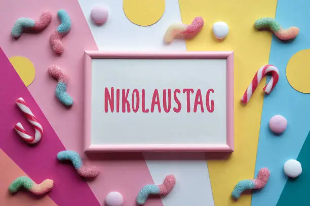 Text Nikolaustag in pink frame means Day of St. Nicholas. Large heart lolly pop, assorted sweets, chocolates, confectionery on multicolor layered paper. Flat lay, top view. Greeting design for December traditional holiday.
